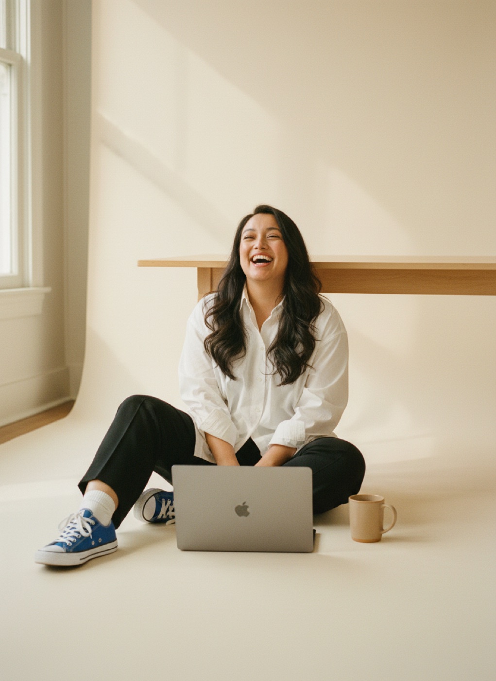 Marielou sitting on a sunlit studio floor laughing, laptop open in front of her, blue sneakers and a coffee mug beside her